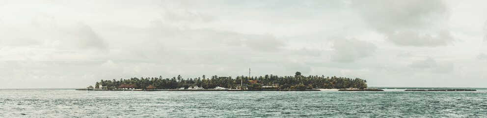 Obraz premium Maldives; Panorama of a Maldivian islet featuring lush green trees, surrounded by the vast calm blue sea, grey clouds in the sky contrast with the overall tranquil scene