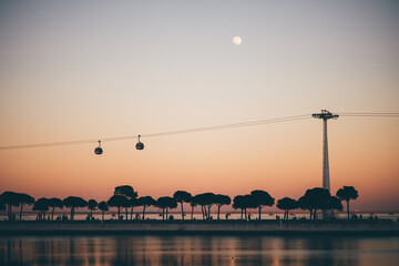 A serene, dreamy low-key view of Lisbon at dusk with a full moon, pink sky sunset, Tejo river, two cable cars, and tree silhouettes by the riverside