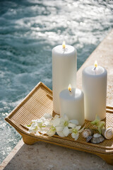 Fototapeta na wymiar Lit pillar candles on tray with white orchids beside pool.