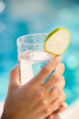 Caucasian woman's hands holding drink glass garnished with lime slice next to pool.