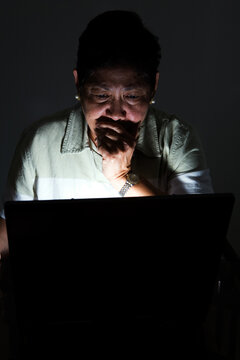 Frightened looking elderly Asian woman looking at the computer screen in the dark with the light coming from the computer