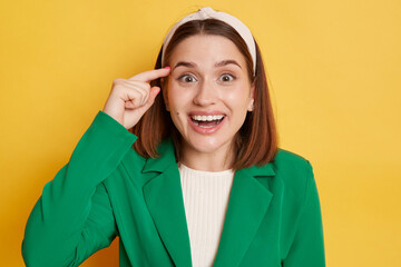 Surprised amazed woman wearing green jacket standing isolated over yellow background looking at...