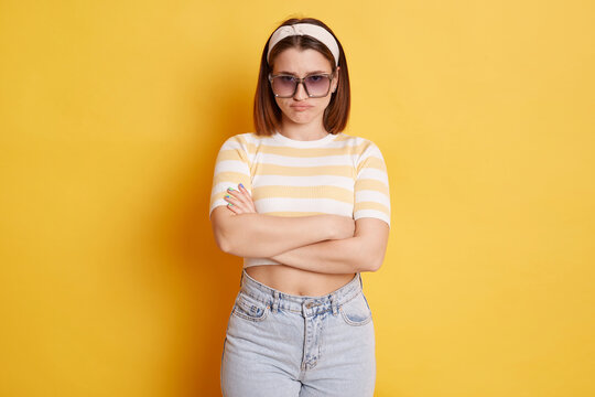 Offended unhappy woman wearing striped T-shirt hair band and sunglasses standing isolated over yellow background standing folded hands expressing sorrow and sadness.