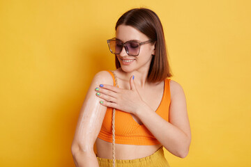 Smiling beautiful woman wearing orange top standing isolated over yellow background applying cream...