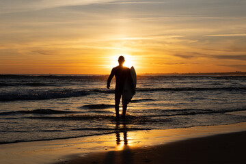Dark silhouette of a man surfer walking out on the seashore holding a surfboard at bright sunset