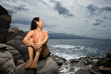 Asian woman sitting on rock by ocean looking over shoulder smiling in Maui, Hawaii