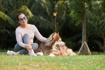 Happy woman sitting on ground in park and stroking her samoyed dog