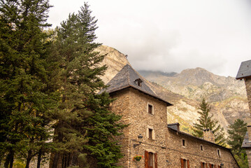 Fototapeta na wymiar Stone house with slate roofs next to large pine trees and an impressive rocky mountain in the background, mountain scenery in the province of Huesca Aragon on a cloudy day. Benasque.