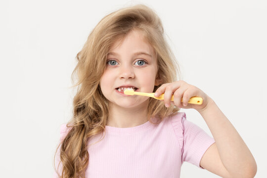 A cute blond child girl on a white background is holding a toothbrush near her face. She smiles broadly and shows the class sign with her finger. Children's dentistry and oral hygiene concept