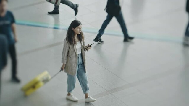 Woman waiting for a flight with suitcase during business trip. Back view of young businesslady in a middle of airport terminal. Girl travelling for a job purpose, waiting for a registration on flight.