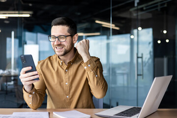 A young businessman works in the office at the table and plays online games on the phone, participates in lotteries and places bets. Happy man celebrates victory, shows victory gesture with his hand