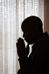 Profile of African-American man silhouetted by a window with his head bowed and his hands in prayer. Vertical shot.