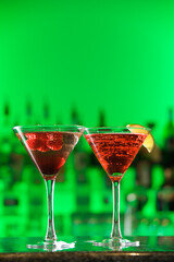 Two cocktails, one with a lemon wedge, sit on a bar counter against a green background. Veritcal shot.