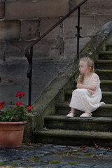 little blond girl in a white dress sits alone on the stairs and looks off to the side, sad mood, dark background, contrast