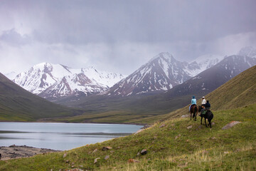 Fototapeta na wymiar Horse trekkers ride their horses past a remote alpine lake in the Tian Shan Mountains of Kyrgyzstan