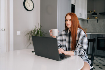 Portrait of thoughtful freelancer female remote working or studying on laptop computer sitting at table, holding in hand coffee cup, thinking looking away. Cute redhead young woman using laptop.