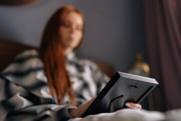 Closeup selective focus shot of sad young woman lying on bed holding photo in frame and crying looking at picture, touching photograph with love. Unhappy female looking to photo with nostalgic feeling