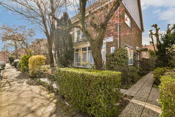 a house with trees in the front yard and bushes growing on the sidewalk next to it, there is a blue sky