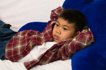Young asian boy laying down on a sofa while looking engrossed in the television