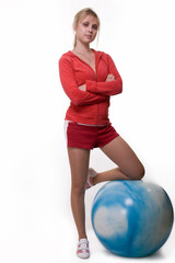 Full body of an attractive blond woman wearing red shorts and sweatshirt standing with one knee on a blue exercise ball over white