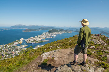 Caucasian tourist wearing a hat standing on top of Sukkertoppen mountain in Ålesund during summer, enjoying the beautiful view of the surrounding islands and ocean. Norway, day, enjoyment