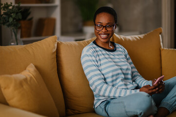 Obraz premium African american woman using a smartphone while sitting on a yellow sofa at home