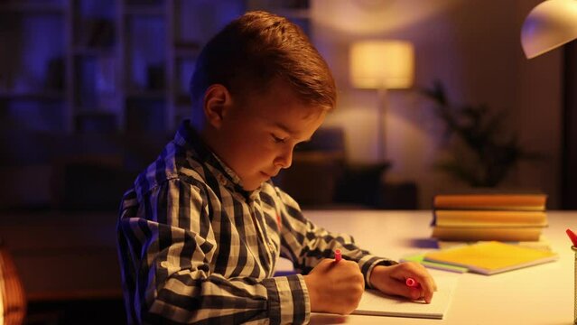 A little brunette boy sits at the table in his room in the evening and draws. A preschooler uses his imagination, creates a picture, dreams of being an artist.