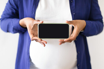 Millennial black woman in domestik clothes with big belly enjoys pregnancy, holds smartphone with empty screen