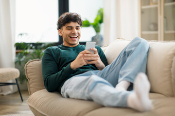 Happy Middle Eastern Young Man Using Cellphone Laughing At Home