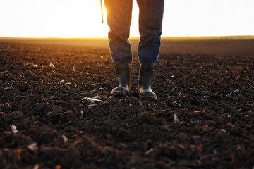Agriculture. Cropped shot of view businessman farmer in rubber boots walks along plowed field with...