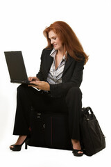 Full body of an attractive red hair woman sitting on a suitcase and typing on a laptop on white
