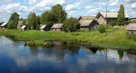 Northern russian village by the river, green grass, blue sky, reflections in water