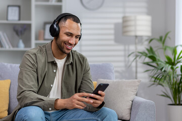 Young hispanic man at home sitting on sofa resting, man with headphones listening online music,...