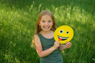 A long-haired blonde girl laughing merrily holds a paper emoticon face in her hand, showing her...