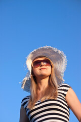 Young smiling blond caucasian white woman with hat and sunglasses. Wearing striped top. Clear blue sky as background. Shot from below. Model looks aside. Copyspace.