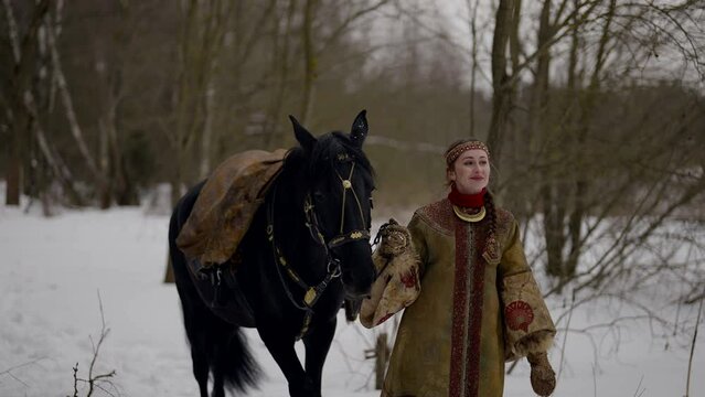 beautiful woman in historical russian gown and black horse walking in winter forest, portrait