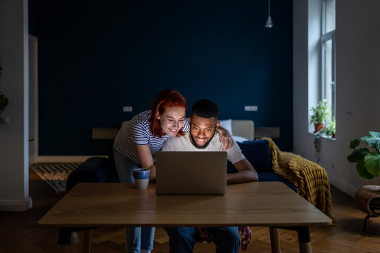 Happy Married Multiracial Couple Using Laptop Together, Planning Summer Vacation Or Shopping Online. White Woman And African American Man Choosing Movie To Watch In Evening At Home, Make A Video Call