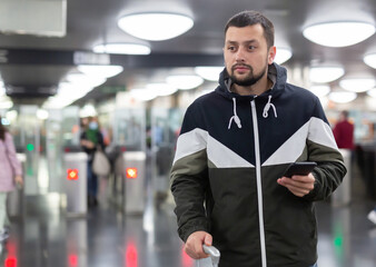 Interested young man browsing messages on phone while waiting for subway train, standing at station..