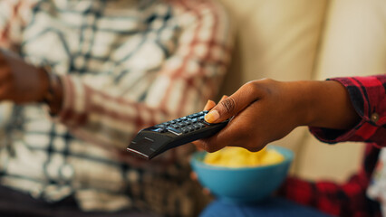 Young girlfriend using tv remote control to watch film with her boyfriend, eating snacks and drinking beer in living room. Modern couple searching for movie on television. Close up.