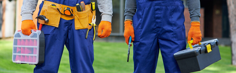 Cropped shot of two male builders in blue overalls carrying toolbox at construction site