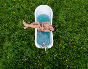 View from above of a young beautiful naked sexy woman, taking outdoors a wellness bath in a blue water bathtub in green nature meadow with yellow buttercups