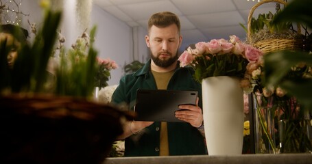 Male florist counts flowers in vase and takes customer order online using digital tablet computer. Small business entrepreneur, owner in flower store. Concept of floral business and entrepreneurship.