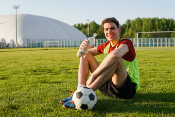 Young man football player resting after training sitting on grass and drinking water from a bottle....