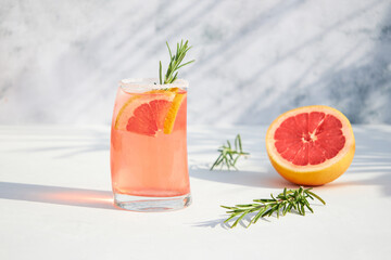 Naklejka na ściany i meble Elegant glass with a fresh, cooling cocktail of fresh grapefruit or red orange with ice cubes and a sprig of rosemary on a gray background on a bright sunny day. Vacation and summer drinks concept.