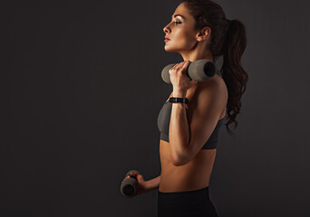 Female sporty muscular young serious woman doing strength workout on the shoulders, biceps and arms in sport bra holding dumbbells on grey background with empty space. Closeup
