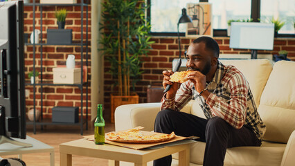 African american guy eating slices of pizza from takeout, drinking bottles of beer and feeling relaxed at television. Cheerful young man enjoying action movie or tv show to binge watch.