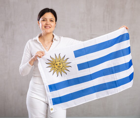 Portrait of young smiling woman in casual clothes posing with flag of Uruguay in studio
