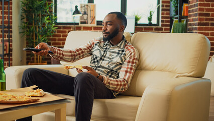 Relaxed guy taking bite of slice of pizza in living room, eating fast food delivery meal and drinking beer from bottle. Young man having fun watching comedy movie on television, eat snacks.