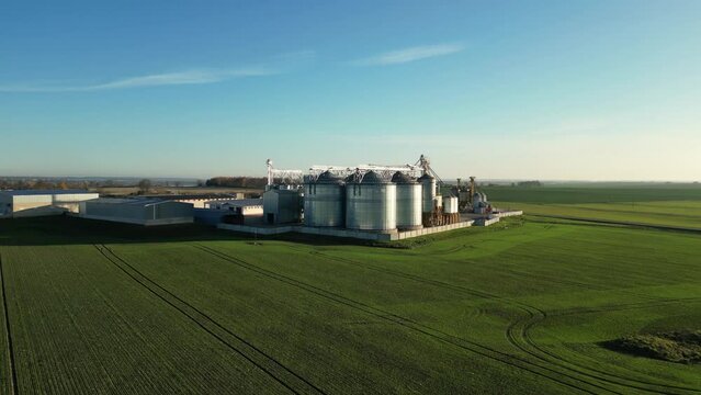 Drone flight over an agricultural field and silver silos at the agro manufacturing plant for processing, drying, cleaning and storage of agricultural products, flour, cereals and legumes.