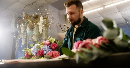 Concentrated male florist wraps beautiful flowers bouquet, uses wrapping paper. Colleague walks at background. Working in flower shop process. Concept of retail floral business and entrepreneurship.